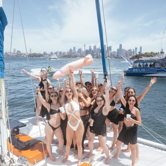 A group of people are celebrating on a luxury yacht hire in Sydney Harbour, enjoying drinks and a large inflatable object against the backdrop of city skyscrapers. The sunny harbor setting is heightened by the presence of another boat nearby.
