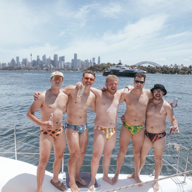 Five men stand on a boat, wearing colorful swim briefs, and posing with arms around each other in front of a city skyline. It looks like they're enjoying an amazing Catamaran party in Sydney!