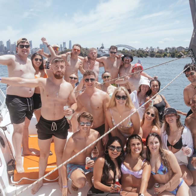 A group of people in swimsuits are gathered on a luxury yacht hire Sydney in a harbor, posing for a photo with the city skyline and a bridge in the background.
