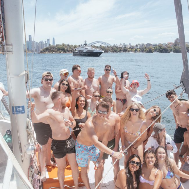 A group of people in swimwear posing for a photo on a boat during a stunning Sydney boat party hire, with a cityscape and bridge in the background on a sunny day.