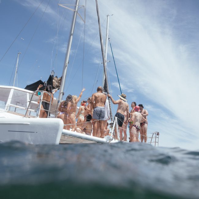 A group of people in swimwear stand and talk on the deck of a white catamaran sailboat on a sunny day with a clear blue sky, enjoying a luxury yacht hire Sydney experience.
