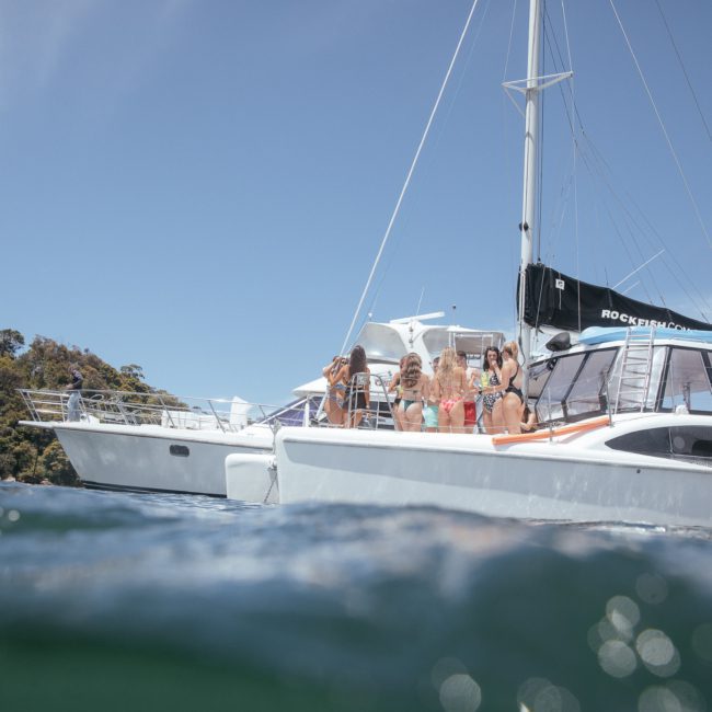 Two boats float on clear water near a forested shoreline. Several people are gathered on the boats, some of whom are standing and others are seated, enjoying the sunny day. Ideal for a private yacht charter Sydney Harbour or corporate boat events Sydney.