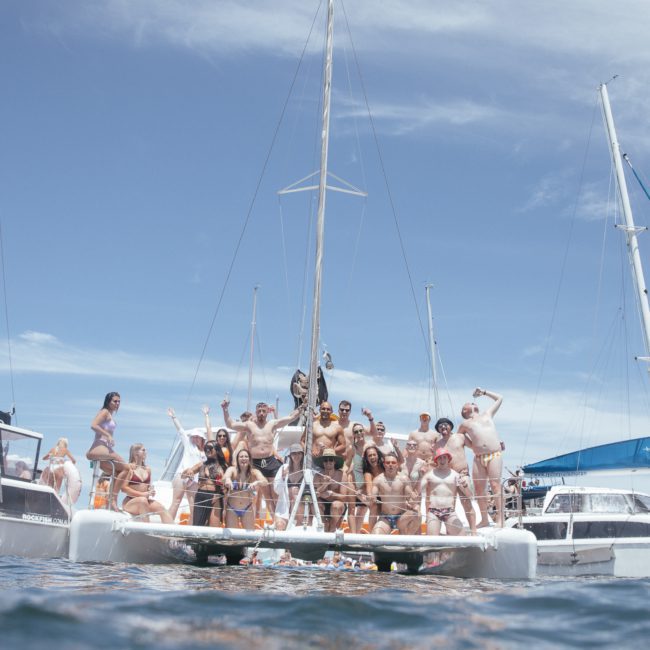 A group of people in swimwear are standing and sitting on a catamaran boat on the water, with other boats nearby under a clear blue sky, enjoying their Catamaran party Sydney.