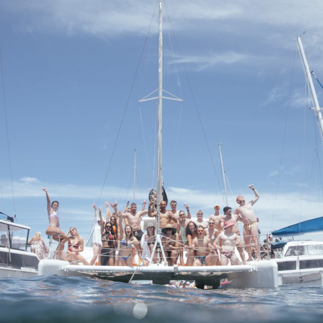 A group of people poses for a photo on the deck of a catamaran during a lively catamaran party in Sydney, with clear skies and other boats visible in the background.