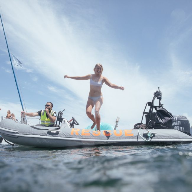 A woman jumps off a small rescue boat into the water while a man in a life jacket looks on, with a large sailboat and Sydney's skyline in the background, enjoying their private yacht charter on Sydney Harbour.