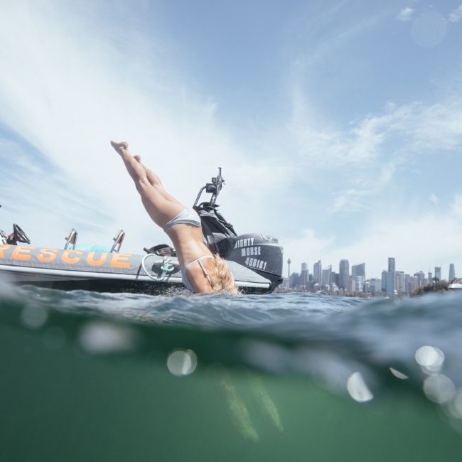 A person in swimwear is diving into the water from the side of a rescue boat with a city skyline and another boat visible in the background during a DJ boat hire Sydney event.