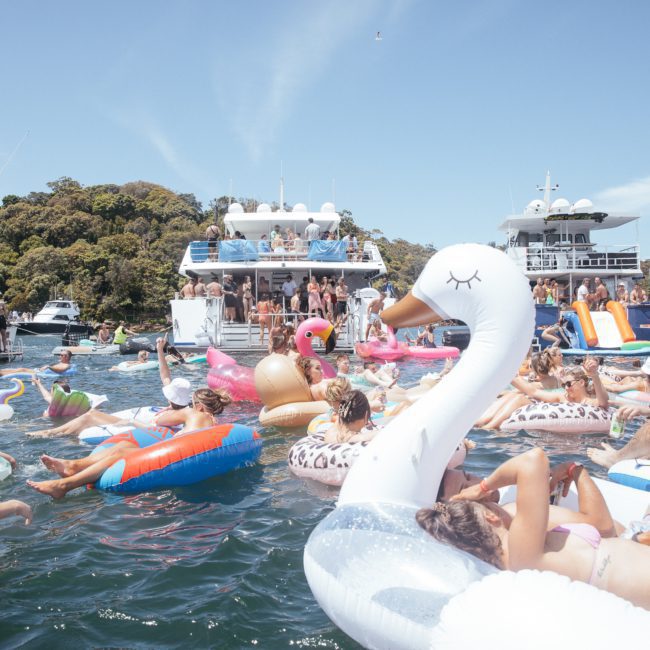 A crowd of people relax on inflatable floats in the water near docked boats on a sunny day, enjoying what feels like a luxury yacht hire Sydney experience.