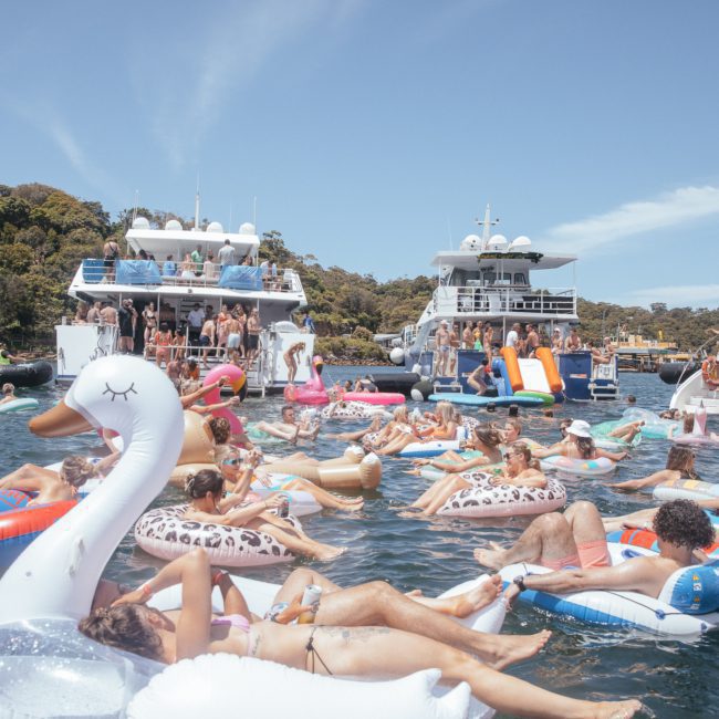 People are relaxing on inflatable floats in the water near a collection of boats on a sunny day, with a lively DJ boat hire Sydney adding to the festive atmosphere.
