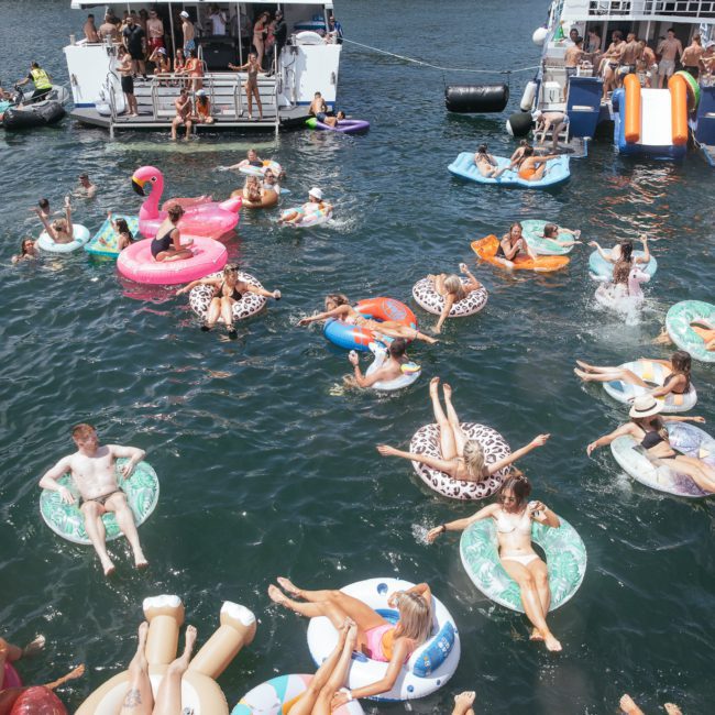 People are relaxing on colorful inflatable pool floats in the water near two anchored boats on a sunny day, enjoying the best of Sydney boat party hire.