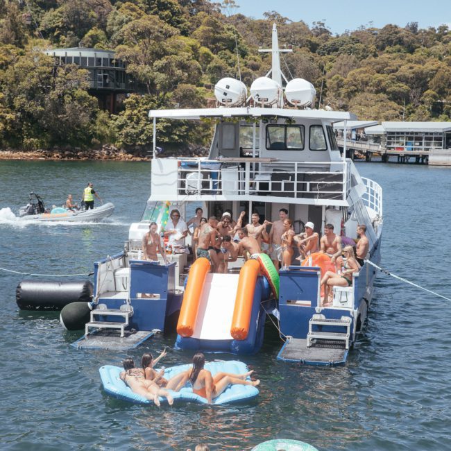 A group of people are gathered on the deck of a catamaran with a large inflatable slide leading into the water. There are inflatables in the water, and other boats are in the background. This scene is perfect for corporate boat events Sydney or a DJ boat hire Sydney experience.