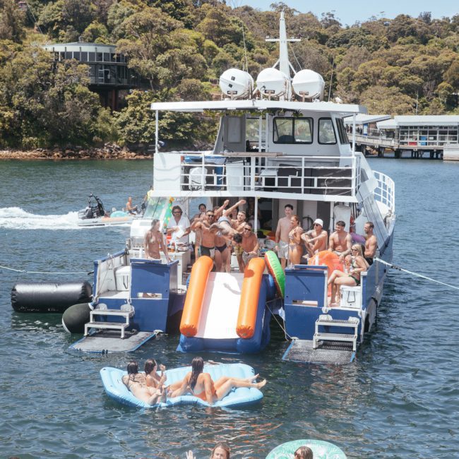People are enjoying a sunny day on a boat with a slide leading into the water. Others are swimming and lounging on inflatable rafts nearby. Trees and buildings are visible in the background, creating the perfect setting for a Sydney boat party hire or private yacht charter in Sydney Harbour.