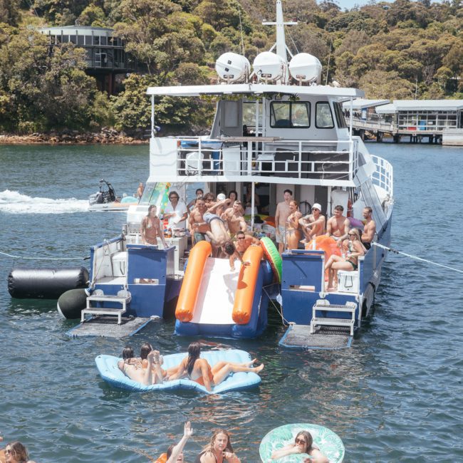 People enjoying a sunny day on a boat, with some using an inflatable slide and others on floating rafts in the water. Trees and a dock are visible in the background—perfect for a Catamaran party in Sydney.
