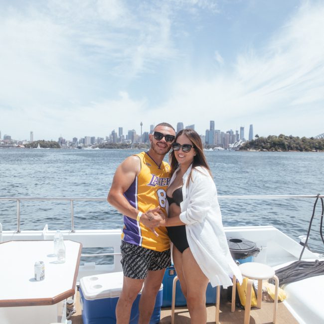 A man and woman standing on a boat deck with a city skyline in the background. The man is wearing a yellow sports jersey and shorts, while the woman is dressed in a black swimsuit and white cover-up. They seem to be enjoying their Luxury yacht hire Sydney experience.