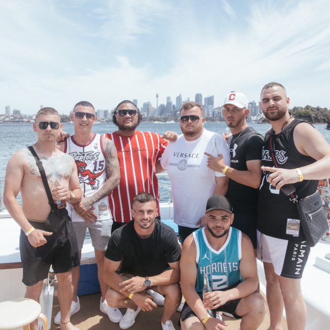 A group of eight men pose for a photo on a luxury yacht hire Sydney with a city skyline in the background. Some are wearing sports jerseys, and several hold drinks. The scene is sunny with a clear sky and calm water, perfect for a memorable Sydney boat party hire.