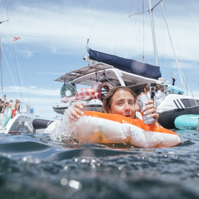 A woman floating in the water on an inflatable holding a can with several boats and people enjoying a Sydney boat party hire in the background.