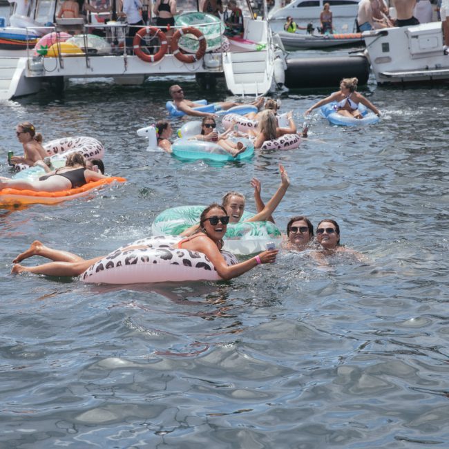 People enjoying a sunny day in the water on inflatable pool floats near boats during a Catamaran party in Sydney, with some waving and smiling at the camera.