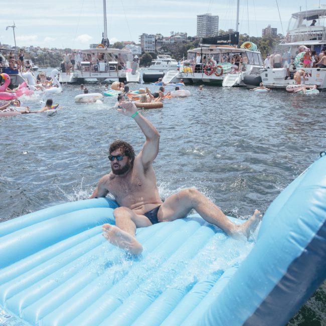Man on a blue inflatable raft in a busy waterway with various people on boats and inflatables in the background, enjoying the vibrant atmosphere of a catamaran party Sydney.
