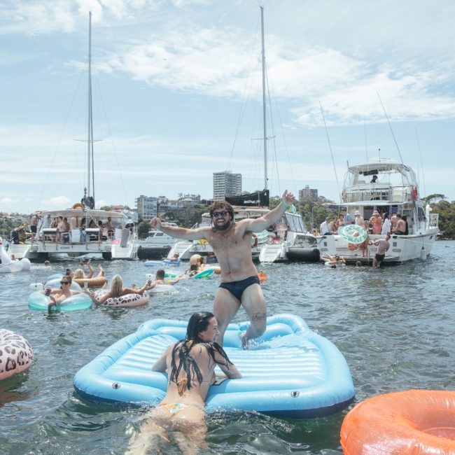 People are enjoying a sunny day in the water on inflatables and boats. A man stands on a blue float, while a woman climbs onto it. Multiple boats, including a luxury yacht hire Sydney, and other people are in the background.