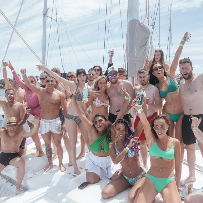 A group of people in swimwear pose and celebrate a Sydney boat party hire on a sailboat under a sunny sky, with the cityscape visible in the background.
