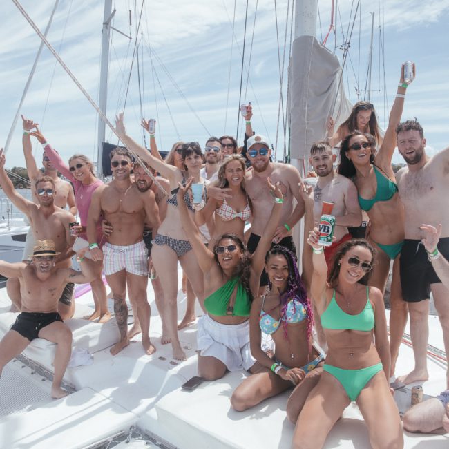 A group of people in swimwear are posing and celebrating on a private yacht charter Sydney Harbour, with some holding drinks. The background shows other boats and a city skyline under a partly cloudy sky.