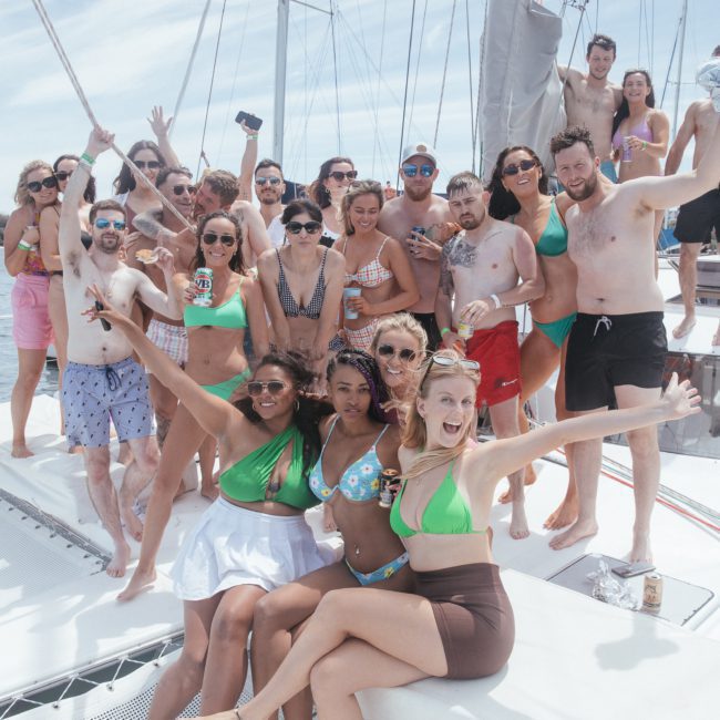A group of people in swimwear are gathered on a boat, smiling and posing for the camera. The background shows a water body and a cityscape of Sydney Harbour, perfect for a private yacht charter or any corporate boat event.