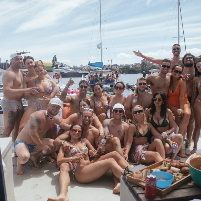 A group of people in swimwear are posing together on a luxury yacht hire Sydney at a marina on a sunny day. There are drinks, food, and other boats in the background.
