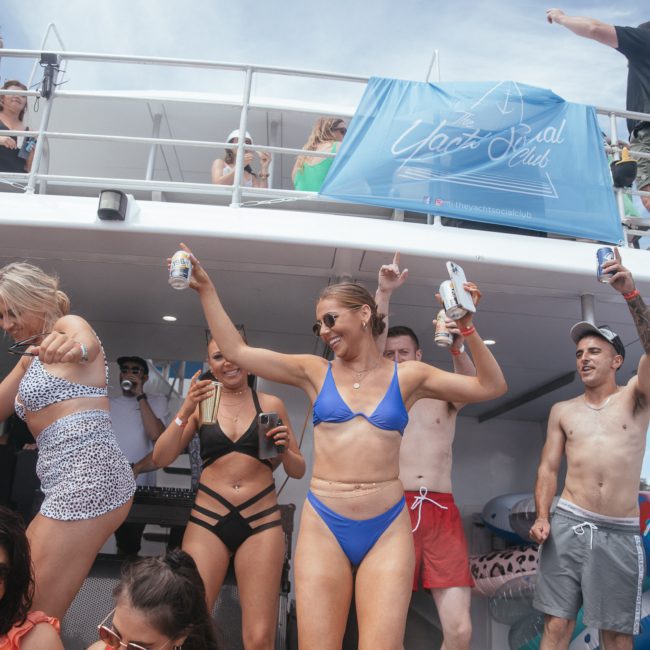 A group of people in swimsuits are partying on a private yacht charter Sydney Harbour, holding cans and drinks. Some are dancing, and others are relaxing on the deck.