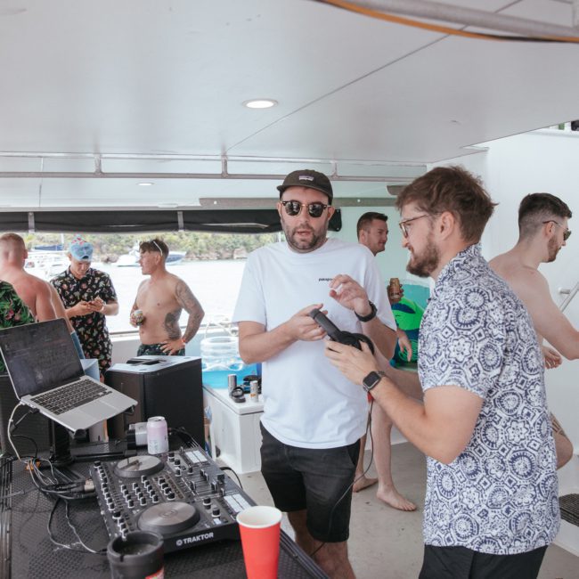 Several people gather on a luxury yacht hire in Sydney Harbour. Two men in the foreground discuss something while one holds a camera. A DJ setup with equipment, including a laptop and speakers, is visible on a counter nearby, creating the perfect ambiance for a memorable Sydney boat party hire.