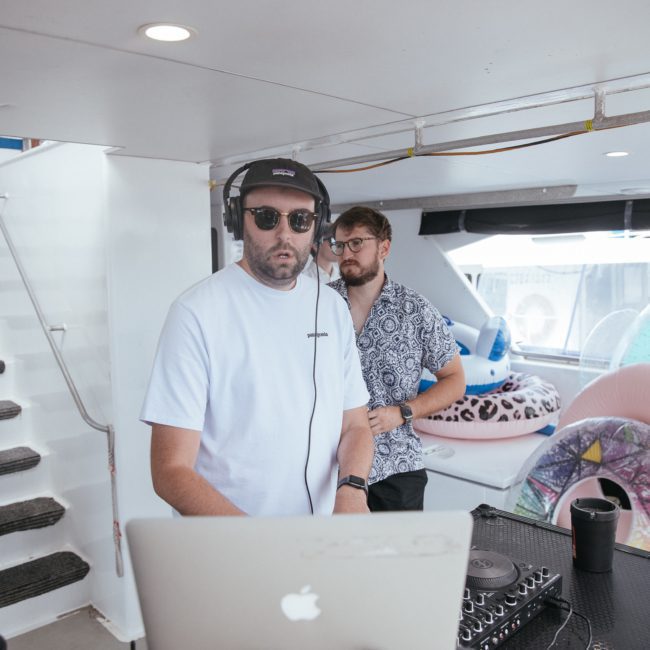 Two men stand behind DJ equipment on a boat. One is wearing headphones and sunglasses while operating the DJ equipment, and the other is standing beside him. Inflatable pool toys are visible in the background during a lively Sydney boat party hire.