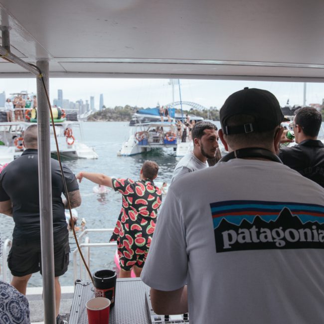 A lively boat party with people socializing and dancing. A person in a "Patagonia" shirt is in the foreground, facing away. Several boats are visible on the water in the background, adding to the vibrant atmosphere of this Corporate boat event Sydney.