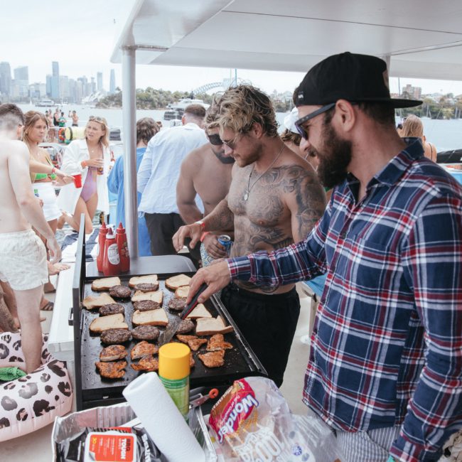 A group of people are grilling food on a boat. One person in a checked shirt and cap is flipping burgers, while another bearded person oversees. Others are relaxing and socializing in the background, enjoying a lively Sydney boat party hire.