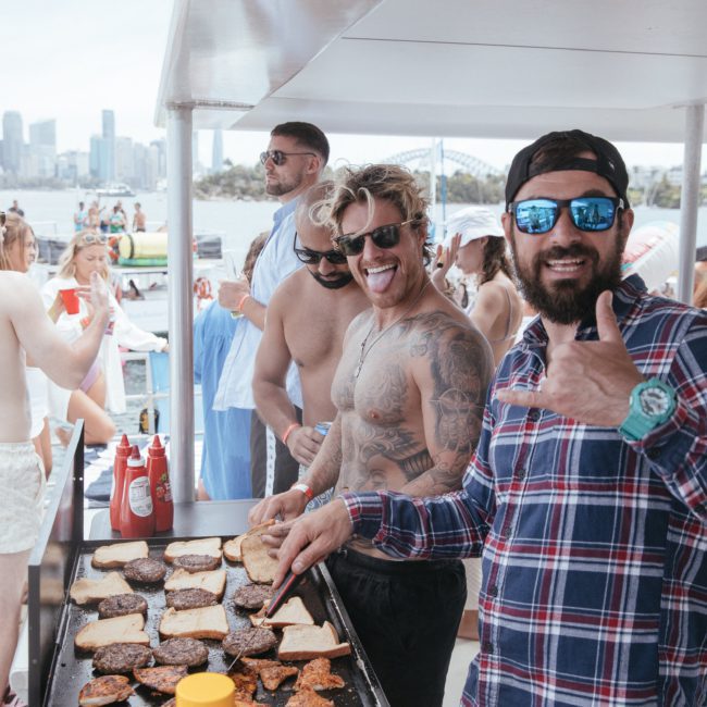 A group of people cooks and prepares food on a grill on a boat with a cityscape in the background. One person in sunglasses and a plaid shirt smiles and gives a thumbs up, capturing the spirit of corporate boat events Sydney.