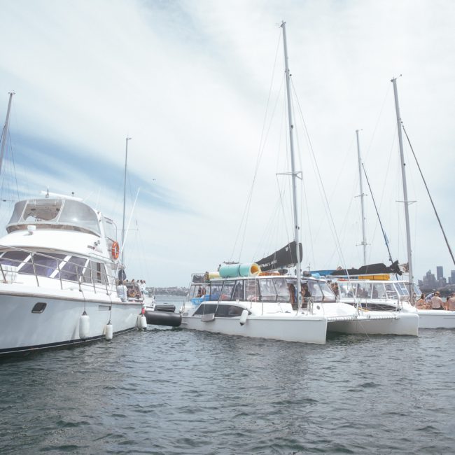 Several sailboats, including a private yacht charter on Sydney Harbour, are anchored together on a calm body of water with city buildings visible in the background under a partly cloudy sky.