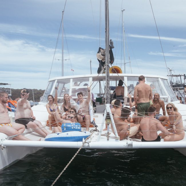 A group of people in swimsuits are gathered on the deck of a white catamaran boat for a Sydney boat party hire in a marina on a sunny day.