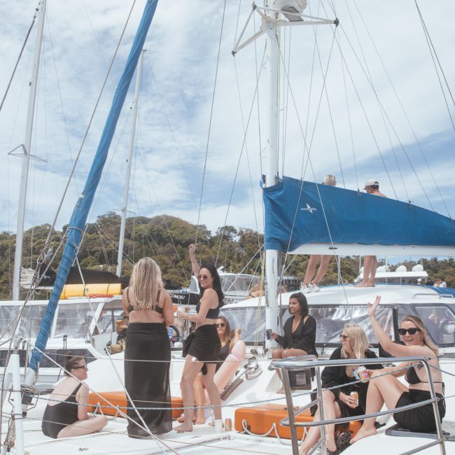 A group of people in black clothing is gathered on a white sailboat in a marina under a clear sky, enjoying one of the popular Corporate boat events Sydney. Trees and multiple boats are visible in the background.
