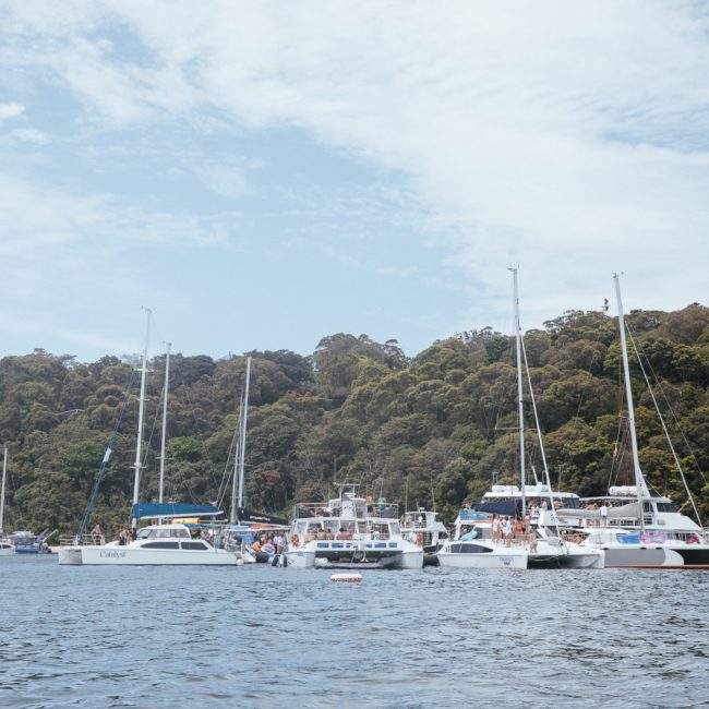 Boats and yachts are docked on calm water near a tree-covered hillside under a lightly cloudy sky, perfect for a private yacht charter in Sydney Harbour.