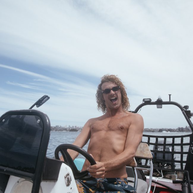 A shirtless man with long curly hair drives a small boat on open water, smiling and looking enthusiastic. The sky is partly cloudy, and a city skyline is visible in the distance, perfect for considering a catamaran party in Sydney.