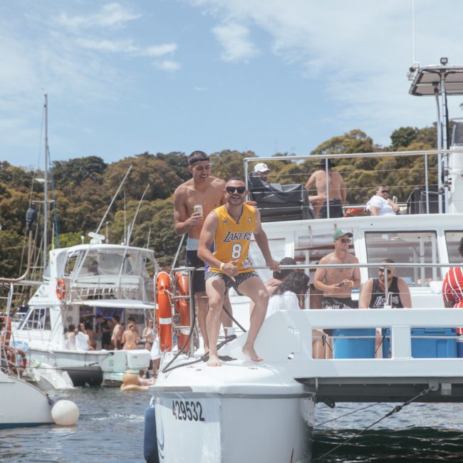 People are gathered on several boats on a sunny day. One man is sitting on the edge of a boat, wearing a yellow sports jersey, while others are standing or sitting around him, enjoying their time. It's the perfect setting for a Sydney boat party hire or luxury yacht hire in Sydney Harbour.