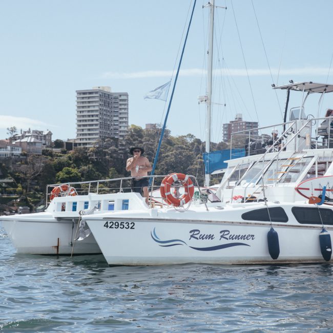 A man stands on the deck of a boat named "Rum Runner" in a marina, with high-rise buildings and trees visible in the background, perfect for a Sydney boat party hire.