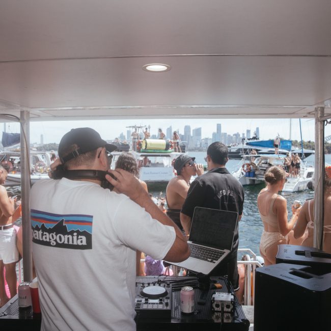 A DJ wearing a white Patagonia shirt spins tunes on a boat using a laptop and DJ equipment. Partygoers in swimsuits dance and socialize around him, creating an energetic vibe. Other boats and people on the water are visible in the background under the bright sun.