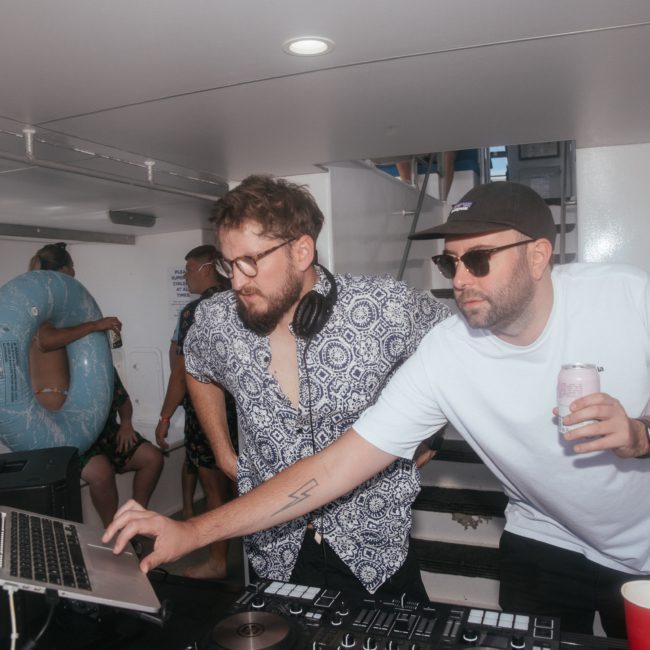 Two men are using DJ equipment and a laptop on a boat. One wears a patterned shirt and headphones, while the other in a white shirt and hat holds a drink. Partygoers and a pool float are visible in the background, enjoying the vibe of this DJ boat hire Sydney experience.
