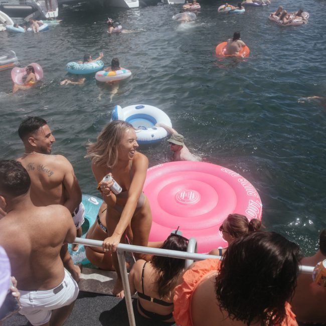 People in swimsuits are enjoying a sunny day on the water with various inflatables. Some are swimming and some are lounging in the water. The atmosphere appears lively and relaxed, perfect for a Sydney boat party hire or private yacht charter on Sydney Harbour.