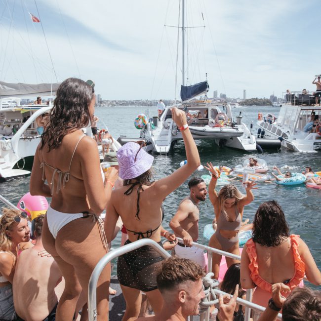 People on a boat enjoying a sunny day, with others swimming and floating in the water near several anchored boats during a lively catamaran party in Sydney.