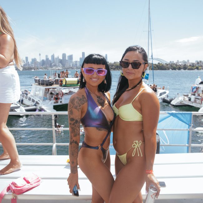 Two women in bikinis stand on a boat with a marina and city skyline in the background. Other people are seen socializing and enjoying the sunny day during a private yacht charter Sydney Harbour event.