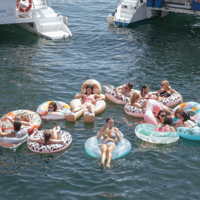 A group of people relaxing on various inflatable floats in water, surrounded by boats, under clear skies, enjoying a private yacht charter on Sydney Harbour.