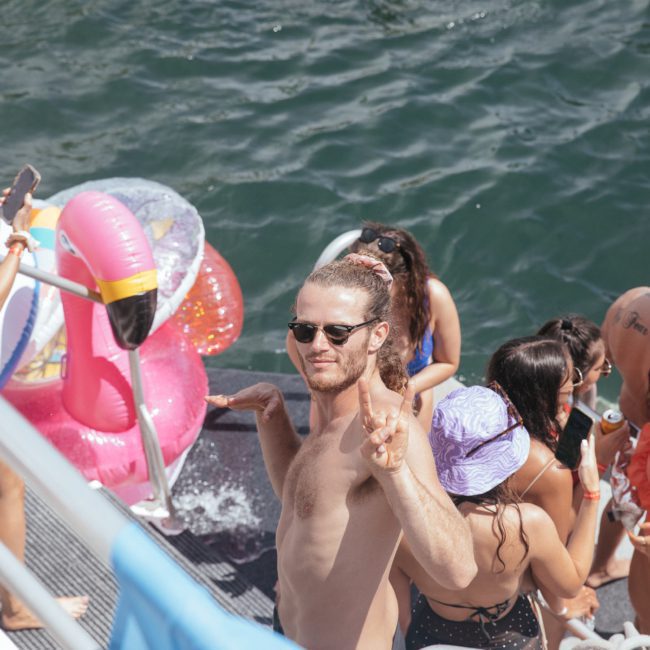 A group of people enjoy a sunny day on a boat with inflatable pool toys in the water. A shirtless man with sunglasses and a ponytail flashes a peace sign while others socialize nearby during a vibrant Sydney boat party hire.