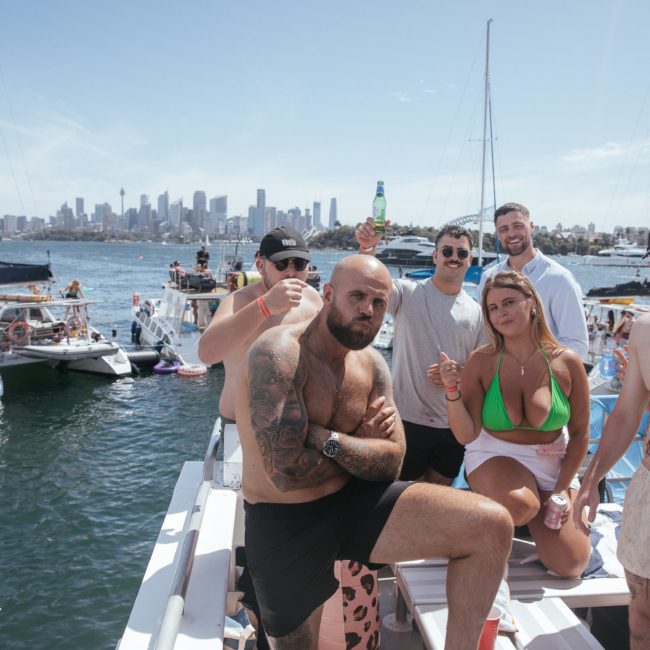 A group of people are socializing on a luxury yacht under sunny weather, with more boats and a city skyline in the background. Some hold drinks and are dressed in swimwear, enjoying a vibrant catamaran party in Sydney.