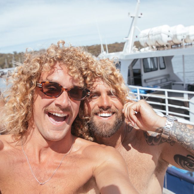 Two shirtless men with curly hair and sunglasses smile and pose for a selfie on a boat deck, with lifeboats and water in the background during their private yacht charter on Sydney Harbour.