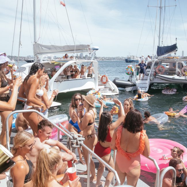 A group of people are gathered on boats and in the water, enjoying a sunny day. Some are on floats, and others are standing or sitting on the boats, holding drinks. The background shows a city skyline—perfect for a lively Sydney boat party hire with friends or colleagues.