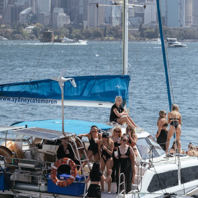 A group of people in swimsuits are gathered on a white catamaran with a “sydneycatamaran.com” banner, enjoying a Catamaran party Sydney, set against a backdrop of city skyscrapers and waterfront.
